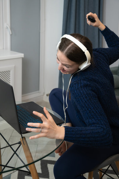 年轻女性戴着耳机，生气地对着笔记本电脑大喊，同时举起手并握着电视遥控器，坐在家里的椅子上。竖版照片图片下载