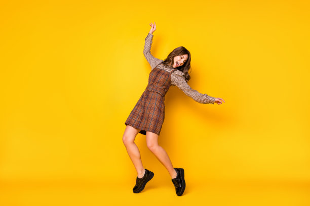 乐观搞笑的棕发女士穿着衬衫裙和运动鞋在鲜艳的黄色背景上跳舞的全身照片图片下载