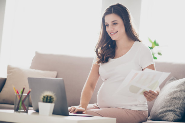 年轻孕妇的侧面照片，开心地微笑，远程工作，家中使用笔记本电脑聊天，报告项目。图片下载