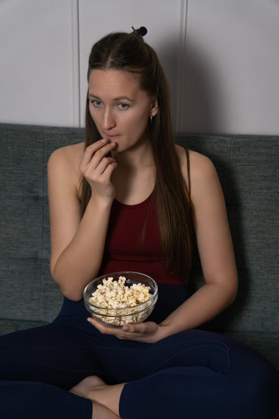 年轻女性坐在客厅的沙发上，正从玻璃碗里吃爆米花，观看一部悬疑或恐怖电影。竖版照片图片下载