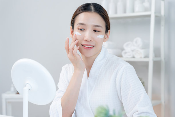 美丽的女性坐在镜子前，健康的面庞正在涂抹面霜和化妆品。皮肤霜、抗皱、婴儿般的面孔、美白、保湿、收缩毛孔精华的广告。图片下载