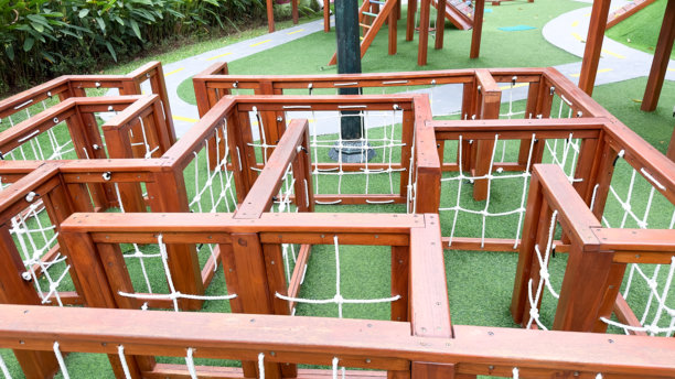 这张图片展示了一个棕色木制迷宫结构，配有白色绳网，置于明亮的绿色人造草坪上。这是一个引人入胜的游乐场设施，旨在让孩子们探索和玩乐。图片下载