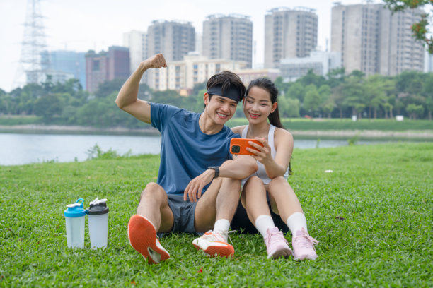 年轻情侣在草地上放松，锻炼后查看手机上的照片并微笑。图片下载