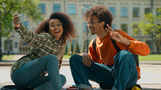 非裔美国学生情侣在城市户外坐着，使用手机自拍，摆姿势面对相机，拍摄大学校园的照片。图片下载