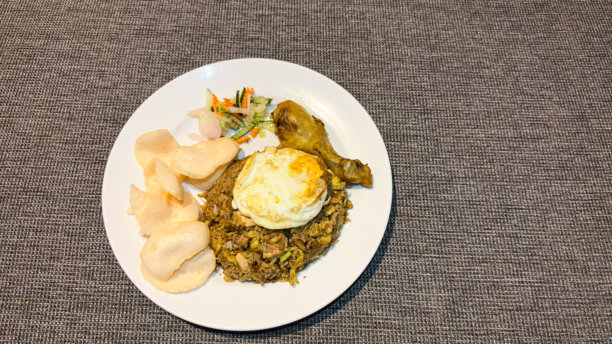 这张图片展示了一道印尼炒饭（Nasi Goreng），配有煎蛋、鸡腿、脆饼和新鲜腌制蔬菜，全部盛放在白色盘子里。图片下载