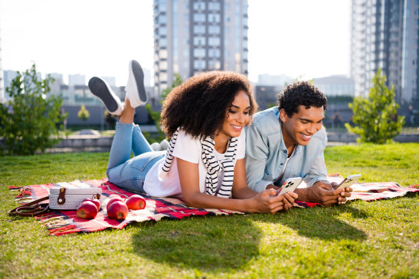 两个兴高采烈的人躺在舒适的毯子草坪上用智能手机阳光休闲公园外面的全尺寸照片图片下载