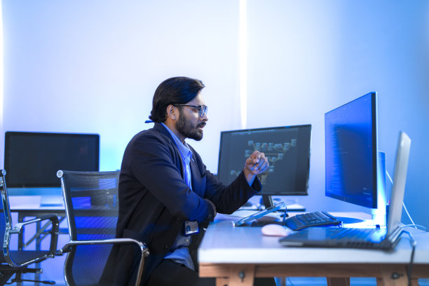 一位专注的印度金融分析师或软件开发人员在现代办公室加班，监控多个屏幕上的数据。金融科技、数据分析和网络安全的概念。图片下载
