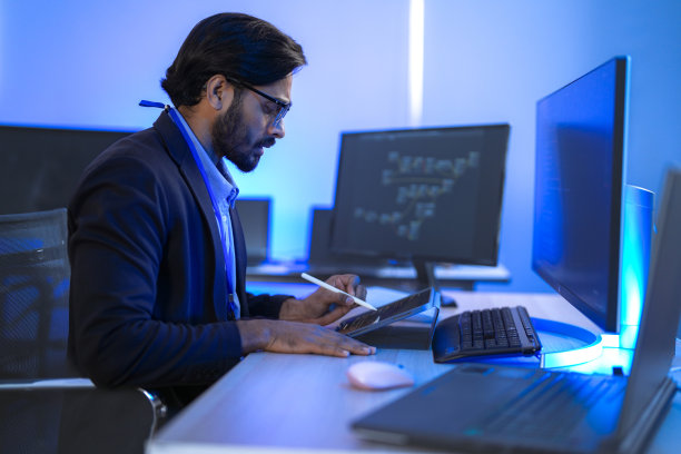 一位专注的印度金融分析师或软件开发人员在现代办公室加班，监控多个屏幕上的数据。金融科技、数据分析和网络安全的概念。图片下载