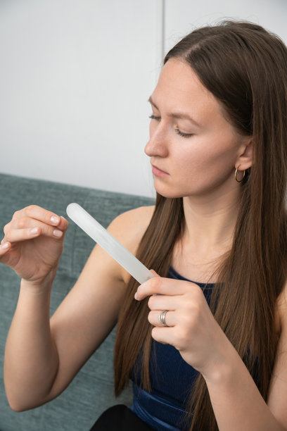 专注的年轻女性坐在舒适的沙发上，使用玻璃指甲锉修整指甲，在家中进行美容护理。竖版照片图片下载