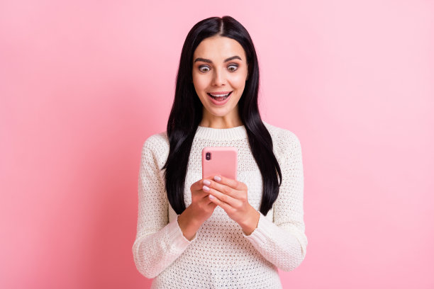 年轻快乐兴奋惊讶的女孩博主在粉色背景下使用智能手机的照片图片下载