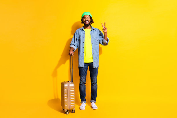 年轻积极微笑的非洲裔男子在黄色背景下展示V字手势，手持行李的全身照片图片下载