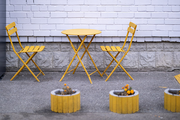 两把黄色金属椅子和一张配套的圆桌放在沥青上，靠在白色砖墙旁，旁边有三个装满五彩花卉的黄色桶形花盆。图片下载