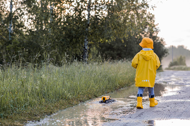 一个穿着黄色橡胶靴和雨衣的男孩在雨后跳过水坑，玩得开心，玩着遥控车。夏季和秋季假期。快乐的童年。图片下载