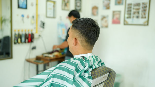 一位发型时尚的男士坐在理发椅上，穿着条纹斗篷。理发师在背景中工作，周围是各种工具和墙上的相框照片，营造出活泼的氛围。图片下载