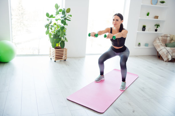 全身照片，漂亮的瘦人双手握着哑铃，蹲在室内粉色健身垫上。图片下载