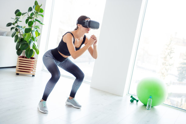 全身照片，快乐、微笑、积极、迷人的运动女孩在家中戴着虚拟现实眼镜进行深蹲锻炼。图片下载