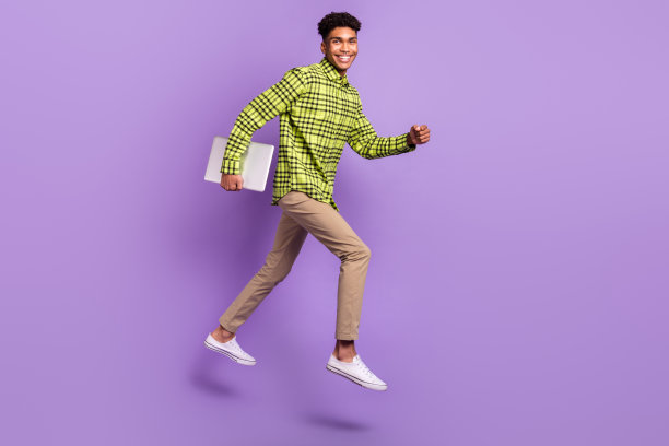 深色皮肤的棕发男性全身侧面照片，跳跃着向空旷的空间伸出手，手中握着笔记本电脑，背景为紫色。图片下载