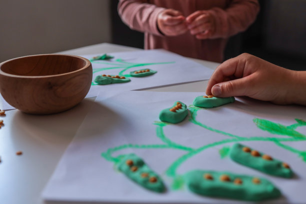 孩子们用粘土和扁豆创造活动，制作手绘植物上的豌豆荚。孩子的手在白色桌子上使用手工材料，散落的扁豆清晰可见。图片下载