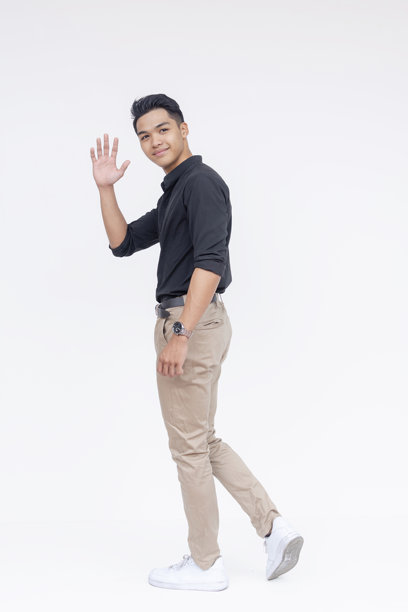 一位友好帅气的菲律宾男子在走开时挥手告别。他穿着黑色的 polo 衫和卡其色裤子。全身照片，背景为白色。图片下载