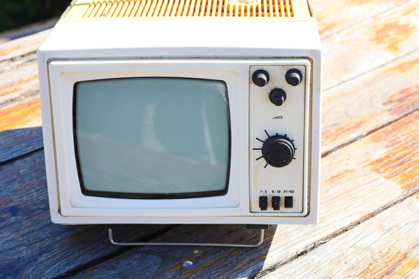 一台旧款便携式电视，白色机身和泛黄的元素，放置在一块磨损的木质表面上，沐浴在明亮的阳光下。这张图片强调了复古美学和对过去科技的怀旧情感。图片下载