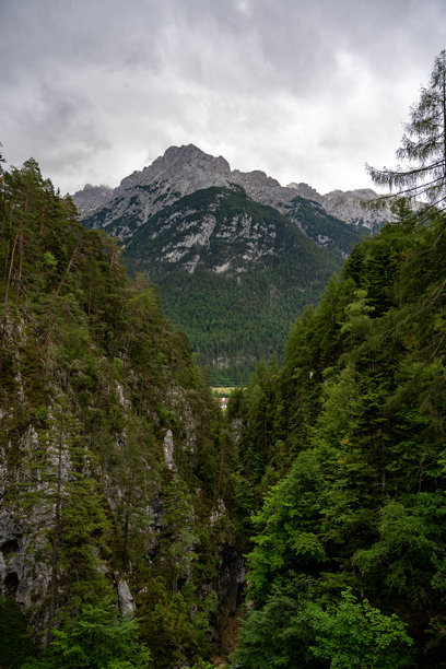 阿尔卑斯山谷景观：雄伟的山峰被郁郁葱葱的森林和崎岖的悬崖环绕，云雾缭绕的天空下 – 自然风光库存照片图片下载