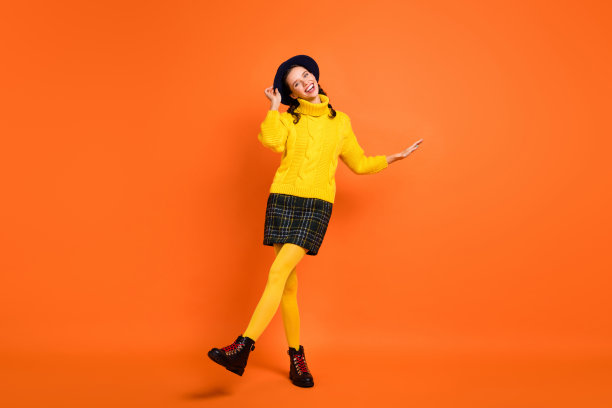 穿着黄色毛衣、帽子、裙子和鞋子的乐观搞笑女孩在橙色背景上跳舞的全身照片图片下载