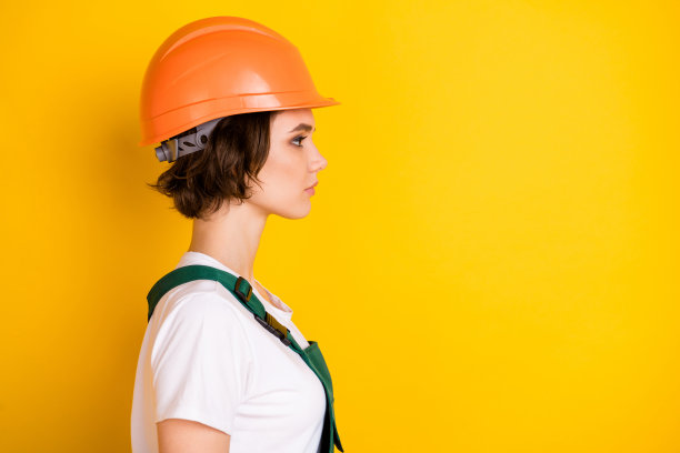 严肃专注的女性建筑工人侧面照片，佩戴安全帽，背景为黄色，留有空白空间。图片下载