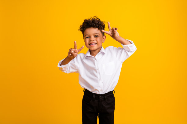 侧面照片，展示了一位穿着白衬衫、留着卷发的可爱时尚男孩，做出V字手势，背景为黄色。图片下载