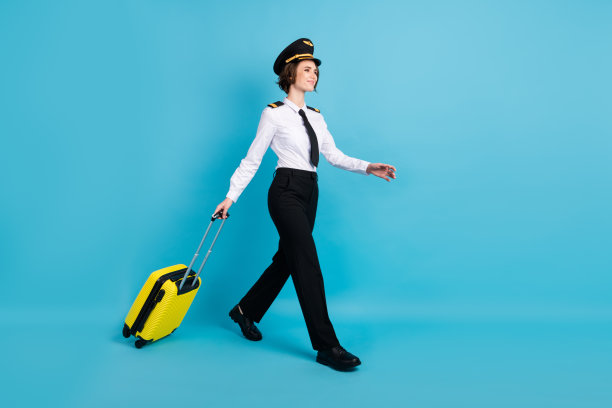 乐观的棕发女士穿着飞行员制服，手提包，走向空旷的地方，背景为蓝色，展示全身侧面照片。图片下载