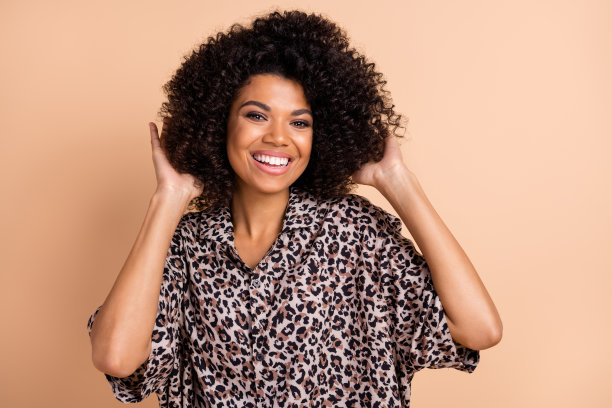 穿着印花衬衫的卷发女性在米色背景前触摸头发，开心地笑着的照片肖像图片下载