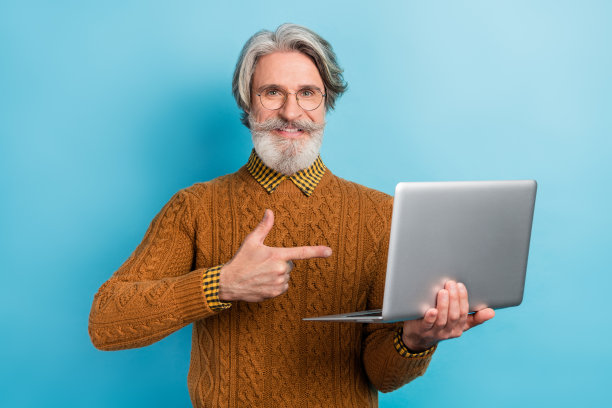 老年商人微笑着指着笔记本电脑的照片肖像，背景为鲜艳的蓝色。图片下载
