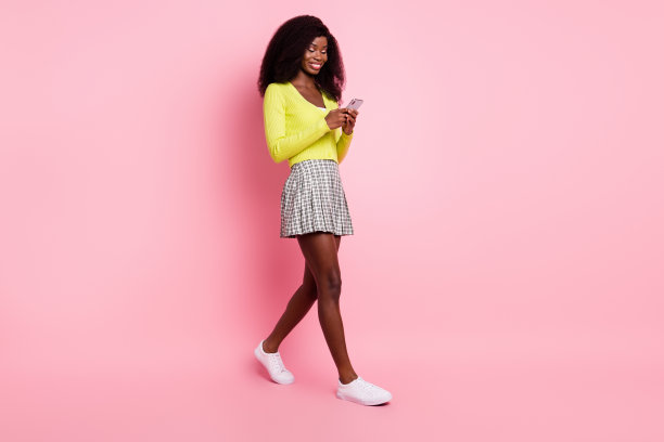 迷人的深色皮肤博主在粉色背景上行走，正在打字发布全身侧面照片图片下载