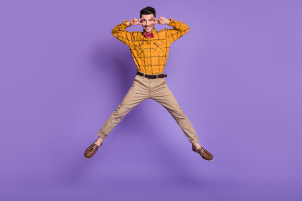 穿着格子衬衫的可爱时尚年轻男孩在紫色背景下跳跃并做出V字手势遮住眼睛的全身照片图片下载