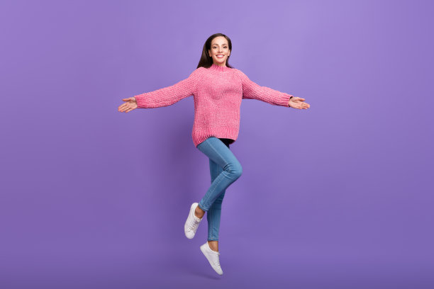 穿着毛衣和牛仔裤的甜美年轻女士在紫色背景上跳跃的全身照片图片下载