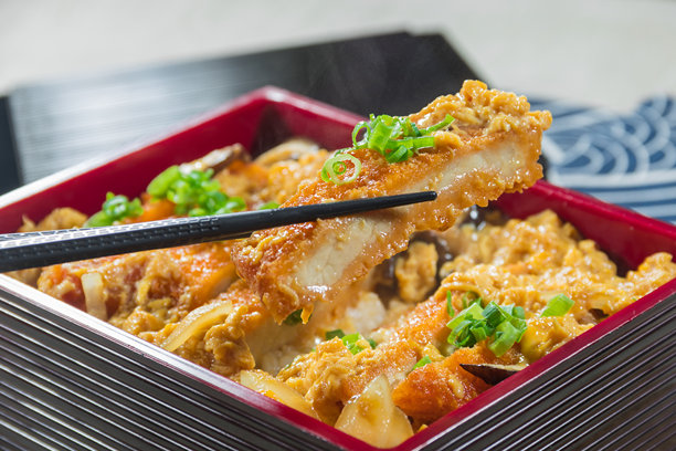 一只漆器日本便当盒里装着一块酥脆的猪排，淋上美味的酱汁，并用葱花装饰，筷子刚刚夹起这道菜，突显了菜单照片中的美食。图片下载