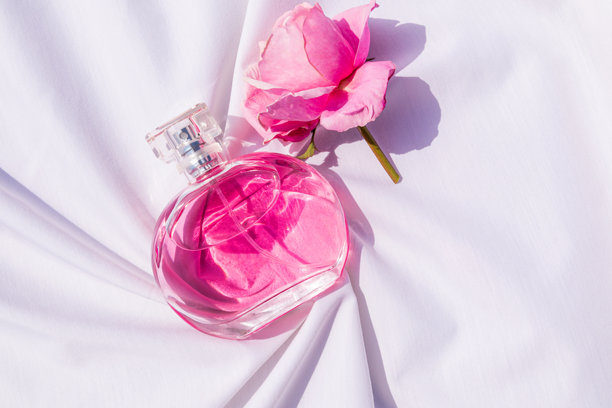 一瓶优雅的女性粉色香水静静地躺在白色缎面织物的柔和褶皱中，旁边是一朵粉色玫瑰花。一个空瓶的展示样本。俯视图。图片下载