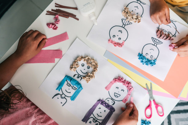 孩子们用皱纹彩纸制作创意纸艺，纸上有卡通面孔和有质感的头发。白色桌子上可见手、胶水和剪刀，以及柔和色调的纸张。图片下载