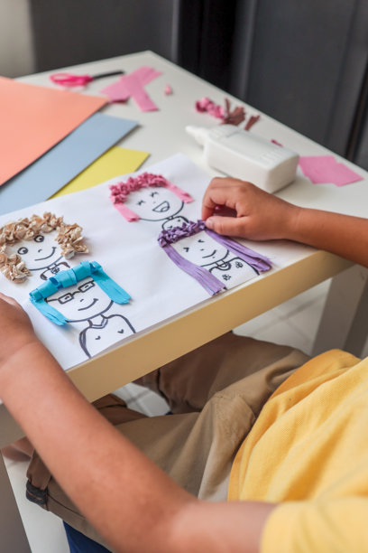 孩子在进行有趣的艺术项目，使用彩色纸发丝粘贴在卡通脸上。桌子上是手工拼贴活动的特写，旁边有胶水、剪刀和粉彩纸。图片下载