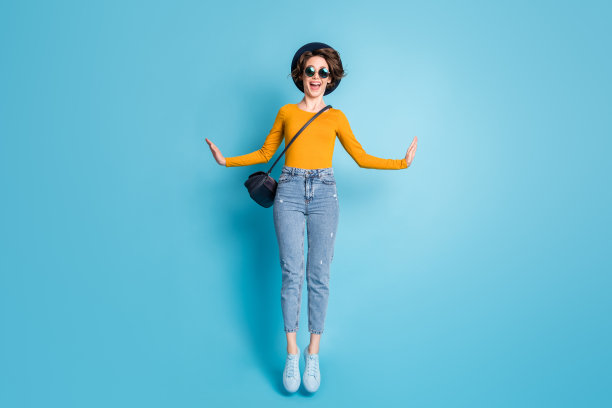 女孩在鲜艳的蓝色背景下，戴着太阳镜和帽子，微笑着跳得很高的全身照片图片下载