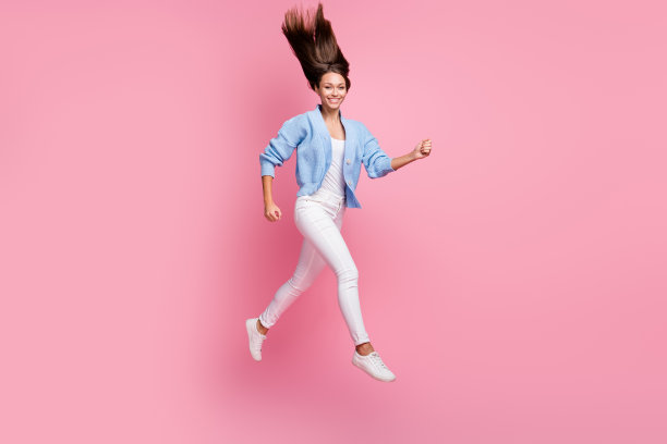 年轻女士全身侧面照片，奔跑跳跃，穿着休闲服，粉色背景留白。图片下载