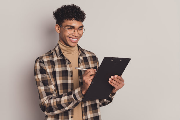 男孩在灰色背景下微笑着，手持夹板进行考试测试的照片肖像。图片下载