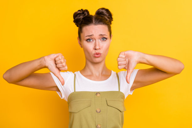 沮丧女孩的照片，做出两个大拇指向下的表情，穿着绿色工装，背景为黄色。图片下载