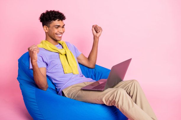 年轻兴奋的黑人男子侧面头像，庆祝胜利，双拳握紧，面向笔记本电脑，背景为粉色。图片下载