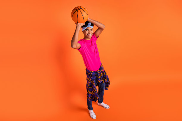 高角度视图的照片，男孩微笑着将球抛向空中，穿着时尚的衣服和头带，背景为鲜艳的橙色。图片下载