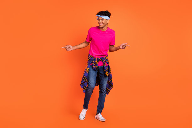 身穿时尚服装、戴着头带的好奇男士在鲜艳的橙色背景下跳舞的全身照片图片下载