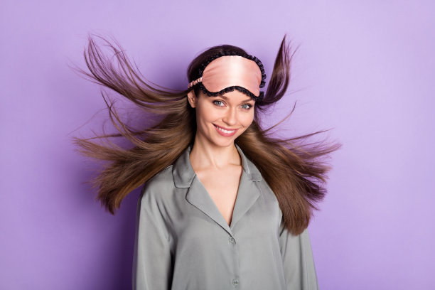 穿着灰色睡衣和睡眠面罩的漂亮女孩的照片肖像，微笑着，愉快地置身于柔和的紫色背景中。图片下载