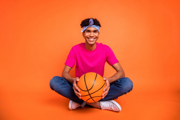 全身照片，男士坐在莲花坐姿，微笑着手持球，穿着粉色T恤，背景为鲜艳的橙色。图片下载