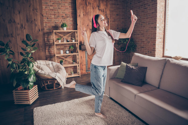 年轻女性全身侧面照片，开心微笑，戴着耳机听音乐，自拍时做出和平手势，背景为家居环境。图片下载