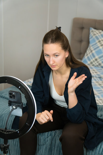 年轻女性网红正在用环形灯和智能手机录制社交媒体视频内容，坐在卧室的床上，手势表达。竖版照片图片下载