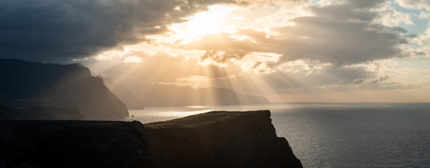 马德拉北海岸的全景照片，拍摄于日落时分。陡峭的悬崖直落大西洋，天空中有戏剧性的云彩和阳光射线。图片下载